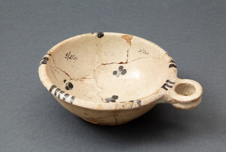 Naczynie ceramiczne. Niewielkie płytkie naczynie zaopatrzone w jedno poziome uszko. Wykonane z jasnej glinki, pokryte od strony wewnętrznej białą polewą, zdobione za pomocą delikatnych czarnych motywów. Jest to lokalny wyrób pomorski z XVI–XVII wieku.