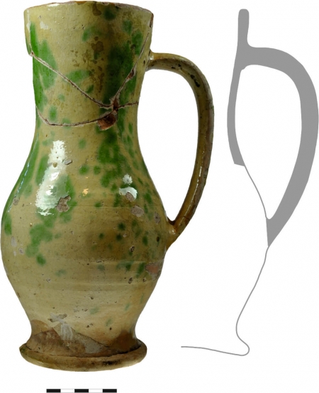 Naczynie z ceramiki ceglastej, szkliwionej, z wyraźnie zaznaczoną stopką, nisko umieszczonym brzuściem i długim, taśmowatym uchem. Jego powierzchnię pokryto najpierw pobiałą, następnie plamiasto nałożoną zieloną angobą, a następnie bezbarwnym szkliwem ołowiowym. XVII wiek (opr. M. Marcinkowski)